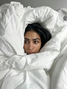 a woman is hiding under a blanket in the middle of a bed with white sheets