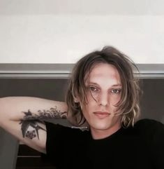 a young man with long hair and tattoos on his arm posing for a photo in front of a window