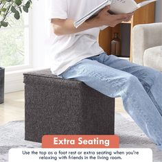 a man sitting on top of a gray ottoman holding a book and looking at it