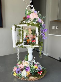 a small white birdhouse with flowers and greenery on it's roof, sitting on a table