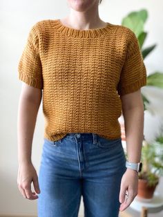 a woman wearing a yellow sweater and blue jeans standing next to a potted plant