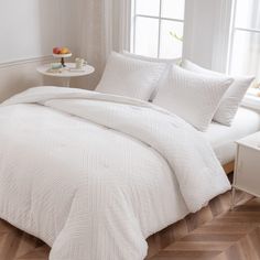 a bed with white comforter and pillows in a room next to a window on a wooden floor