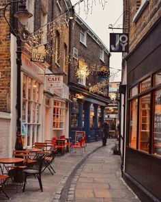 an empty street with tables and chairs on the sidewalk in front of buildings that have lights strung from them