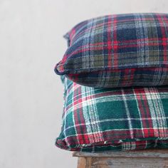 two plaid pillows stacked on top of each other in front of a white wall and wooden stool