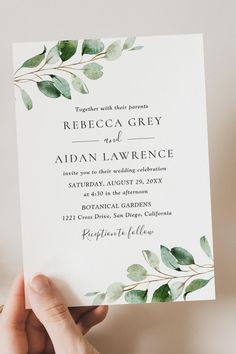 a person holding up a wedding card with greenery on the front and bottom corner