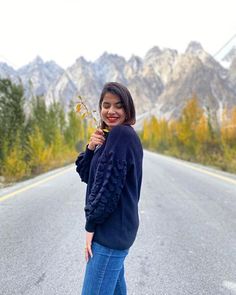 a woman standing on the side of a road holding a flower in one hand and smiling at the camera