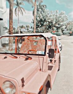 an old pink jeep parked in a parking lot with palm trees and buildings behind it