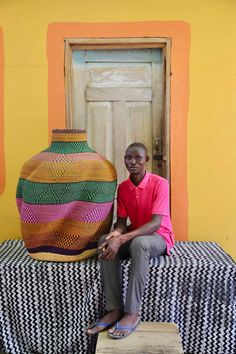 a man sitting on top of a table next to a large vase and ottoman in front of a yellow wall