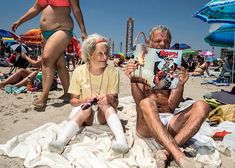 an older woman sitting on the beach reading a book next to a man who is wearing white socks