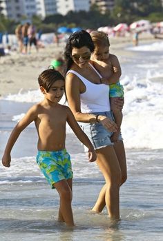 a woman and two children are walking in the water at the beach with buildings in the background