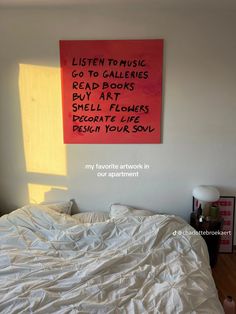 a bed with a white comforter and a red sign on the wall above it that says listen to music go to gallerys read books buy art