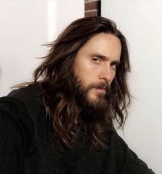 a man with long hair and beard sitting in front of a white wall wearing a black shirt