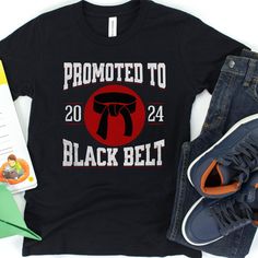 Celebrate a milestone in your young martial artist's journey with this personalized "Promoted to Black Belt" t-shirt! 🥋 This custom tee is not only a symbol of hard-earned success but also a keepsake that commemorates their dedication and the year they achieved this impressive rank. Crafted from comfortable materials that stand up to rigorous training sessions or casual wear, this tee is perfect for any occasion. -Personalize with the year of achievement -Durable and comfortable fabric for acti Black Team Spirit T-shirt With Letter Print, Black T-shirt With Name Print For Fans, Black Graphic Tee With Team Name, Black Crew Neck T-shirt For School Spirit, Black Crew Neck T-shirt With Name Print, Sporty Black T-shirt With Custom Print, Black School Spirit T-shirt With Screen Print, Black School Spirit T-shirt For Sports Events, Black School Spirit T-shirt With Name Print