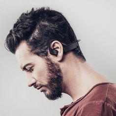 a bearded man wearing an earbud and looking down at his cell phone while standing in front of a white wall