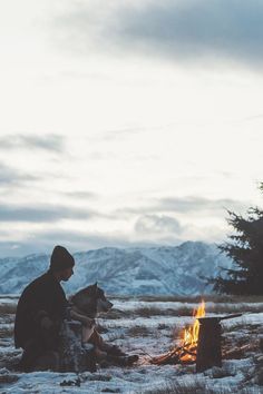 a man sitting in front of a campfire with his dog next to it on a snowy field