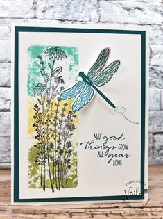 a card with a dragonfly on it and some flowers in the background, sitting on top of a wooden table