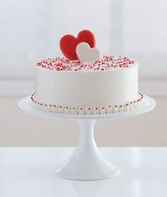 a white cake with red sprinkles and two hearts on top, sitting on a pedestal