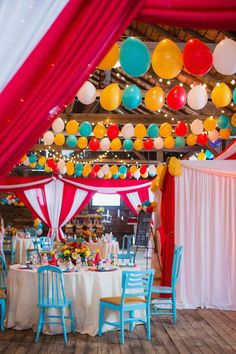 colorful balloons are hanging from the ceiling above tables and chairs in an indoor event venue