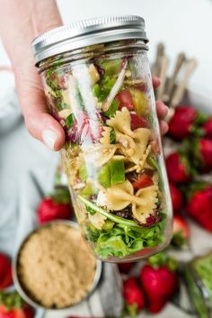 a person holding a mason jar full of pasta salad with dressing and strawberries in the background