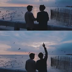 two people standing next to each other in front of the ocean with birds flying around