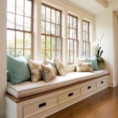 a window seat with many pillows on it in front of a wooden floor and white walls