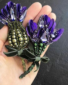 two purple flowers with green leaves and beads on their ends are being held by someone's hand