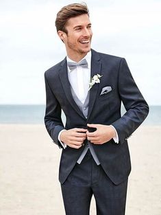 a man in a suit and bow tie standing on the beach with his hands in his pockets