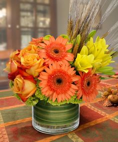 an arrangement of colorful flowers in a vase on a table with grass and corn stalks