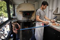 a man standing in front of a pizza oven while people watch from the other side