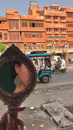 a woman holding up a mirror to her face in front of a busy street with cars and mopeds