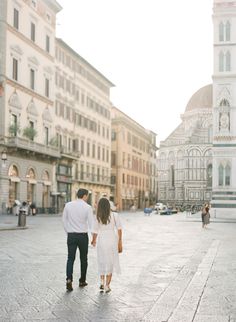 a man and woman holding hands walking down the street with buildings in the back ground