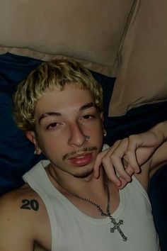 a young man laying on top of a bed wearing a cross necklace and looking at the camera