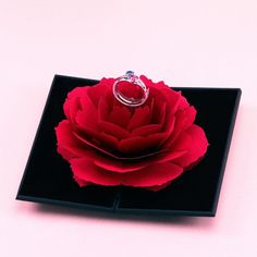 a ring sits on top of a red rose in a black square tray with a white background