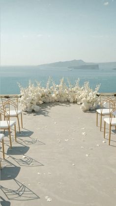 an outdoor ceremony setup with white chairs and flowers on the ground next to the ocean