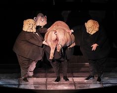 three men in masks holding a pig on stage