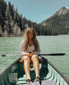 a woman is sitting in a boat on the water with mountains in the background and trees around her