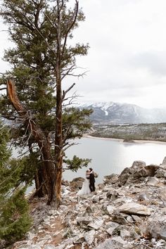 a bride and groom standing on top of a rocky hill next to a large body of water