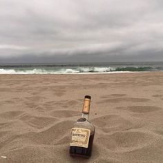 a bottle of liquor sitting on top of a sandy beach next to the ocean under a cloudy sky