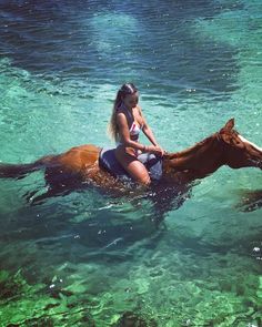 a woman riding on the back of a brown horse in clear blue water with green algae