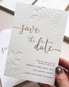 a person holding up some white cards with gold foil on them and the words save the date written in cursive writing