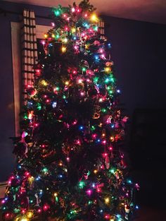 a brightly lit christmas tree in the corner of a room with a window behind it