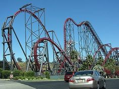 the roller coaster at an amusement park with cars parked in front and on the road