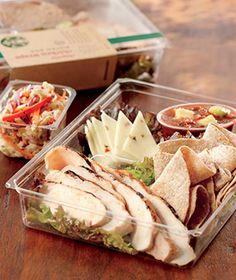 two plastic containers filled with food on top of a wooden table next to a box of chips