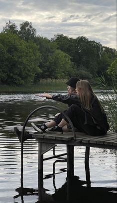 a man and woman sitting on a bench in front of a lake pointing at something