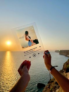 someone holding up a photo with the sun setting in the background and water behind them