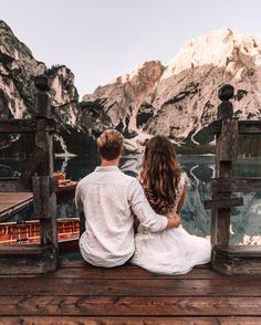 a man and woman sitting on a pier looking at the mountains