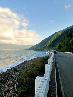 an empty road next to the ocean with cars on it and mountains in the background