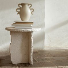 a white vase sitting on top of a table next to a stack of books in front of a wall