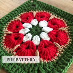 a green and red crocheted square with white flowers on the center is sitting on a wooden surface