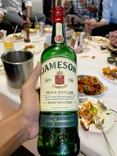 a person holding up a bottle of jameson on a table with other people eating in the background